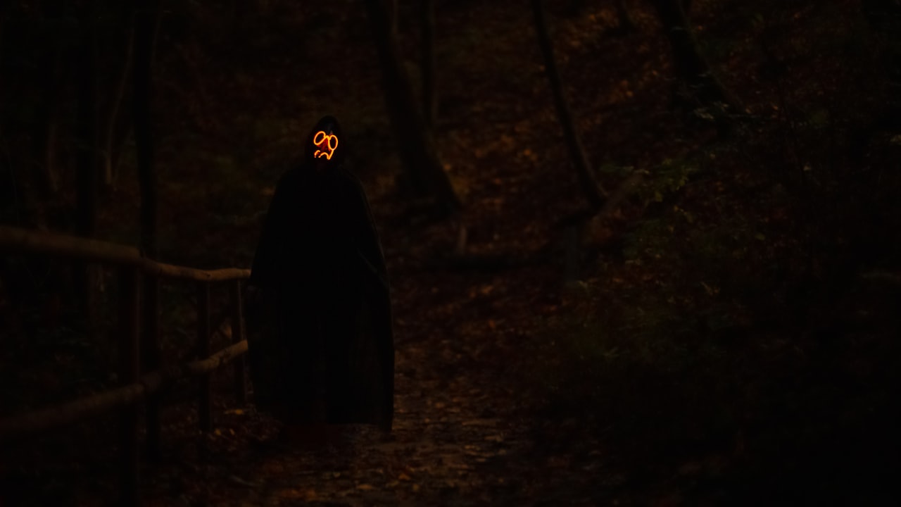 A lone figure in a long black cloak walking into a dark, foggy forest