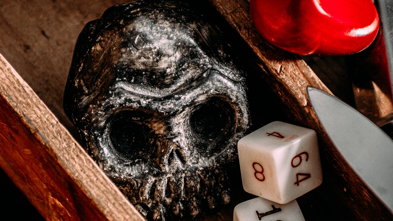 A weathered skull, a polyhedral die with crimson pips, and a folded blade arranged on a dark wooden tabletop